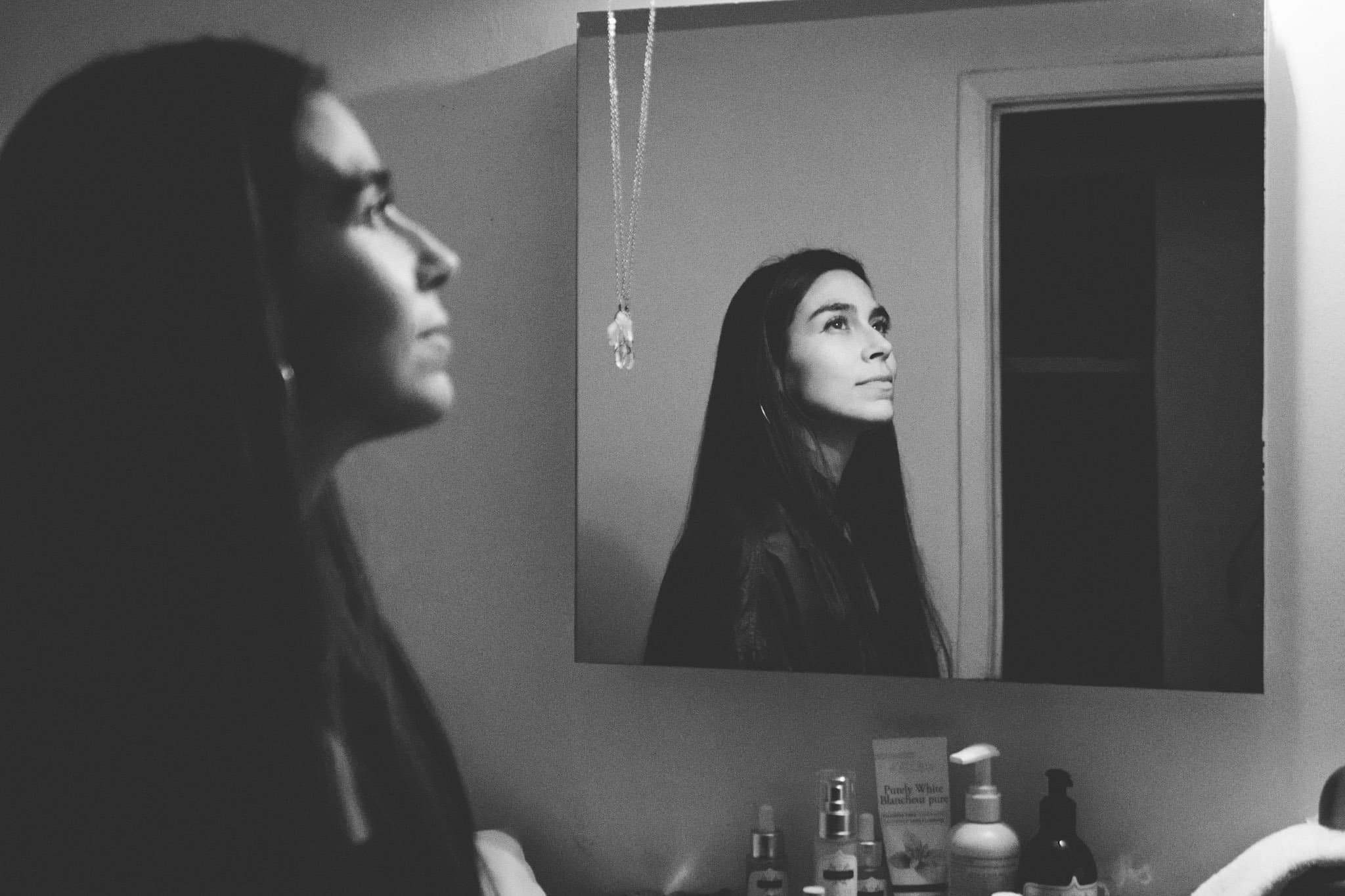 A woman looking at herself in a mirror in a bathroom, with various skincare products on the counter in front of her.